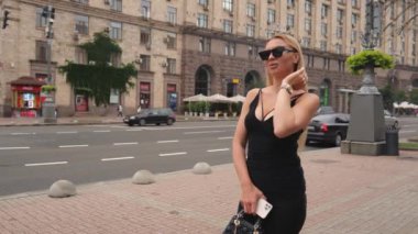 Koyu renk elbiseli ve gözlüklü genç bir kadın ana caddede duruyor ve binaların etrafında arabalar hareket ediyor. Bir kadının yavaş hareket eden portresi. Sarışın kadın.