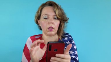 Kırmızı tişörtlü yetişkin kadın mavi arka planda Amerikan bayrağı olan bir cep telefonuna bakıyor. Kadın portresi