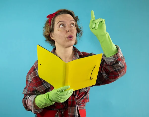 An adult pin-up woman in a plaid shirt holds a yellow book in her hands on a blue background. Woman in green rubber cleaning gloves. Pin-up woman portrait