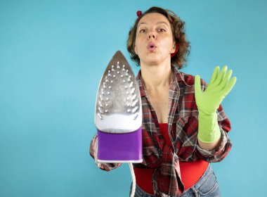 Yetişkin bir kadın mavi arka planda kameraya ütü gösteriyor. Ekose gömlekli bir kadın portresi. Demir kıyafetler.