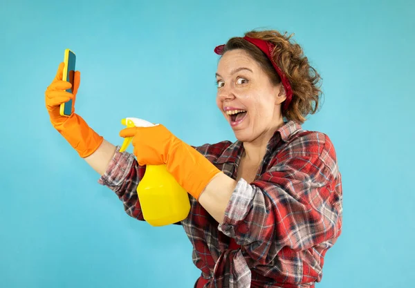 A pin-up cleaning woman in a plaid shirt holds a mobile phone and a spray bottle in her hands on a blue background. Portrait of a cheerful pin-up woman. Adult woman wearing rubber green cleaning gloves