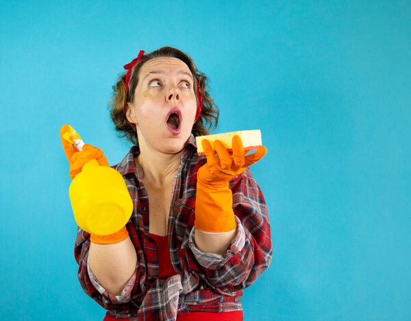 Adult pin-up woman cleaning in a plaid shirt holds a sponge and a yellow spray gun in her hands on an isolated blue background. Portrait of a woman. Woman with open mouth