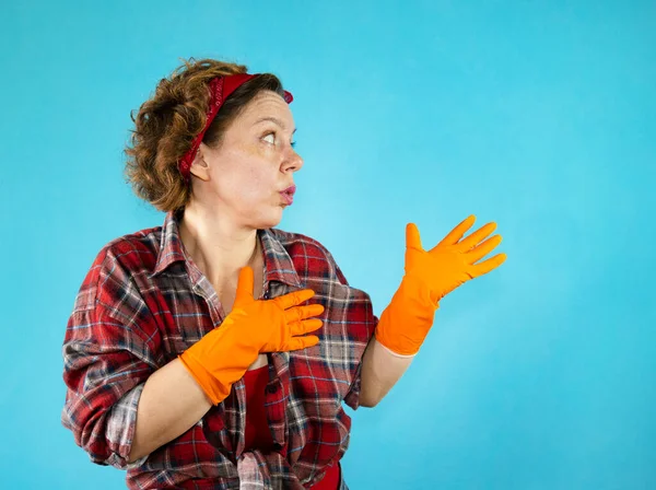 Woman pin-up cleaning in a plaid shirt shows with her hands on an isolated blue background. Cheerful woman. Portrait of woman cleaning. Orange rubber gloves