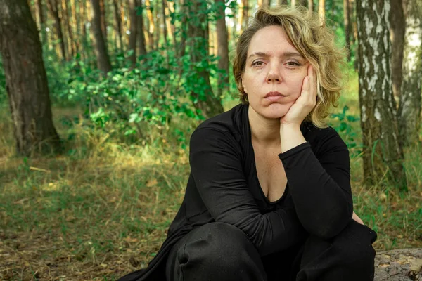 An adult woman in black clothes sits on a log in the forest. Woman in dark clothes
