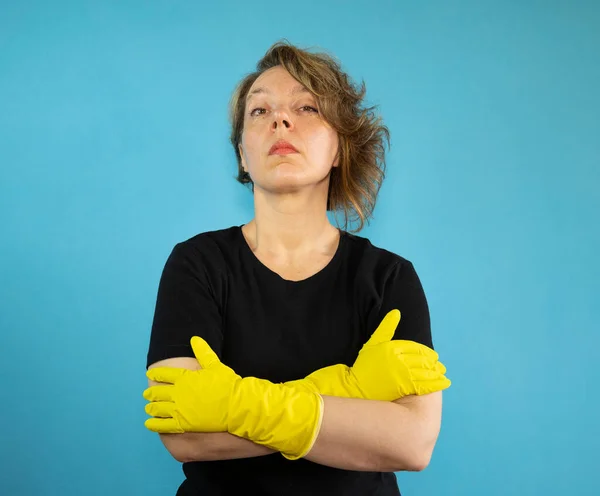 Portrait of a serious cleaning woman in a black t-shirt on an isolated blue background. Woman in yellow rubber gloves for cleaning