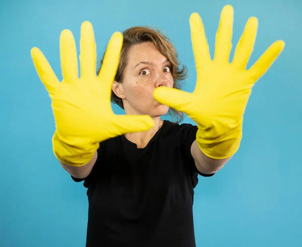 Cheerful cleaning woman shows hands in yellow rubber gloves on a blue isolated background. Portrait of woman cleaning in black t-shirt