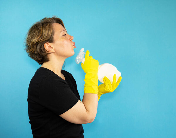 Profile view of an adult woman cleaning in a black t-shirt with a white spray bottle on an isolated blue background. Portrait of woman cleaning in t-shirt