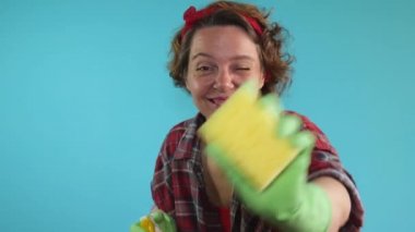 A pin-up woman in a plaid shirt holds a yellow spray bottle and a sponge for washing in her hands. Cheerful cleaning woman in green rubber gloves on a blue isolated background. slow motion