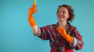 An adult pin-up woman in a plaid shirt and rubber orange gloves takes a selfie on a mobile phone. Portrait of woman cleaning
