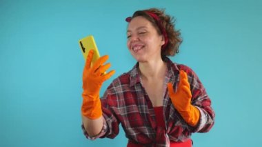 Adult woman cleaning pin-up communicates by video communication through a mobile phone on a blue isolated background. Woman cleaning in rubber orange gloves