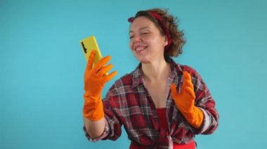 Adult woman cleaning pin-up communicates by video communication through a mobile phone on a blue isolated background. Woman cleaning in rubber orange gloves