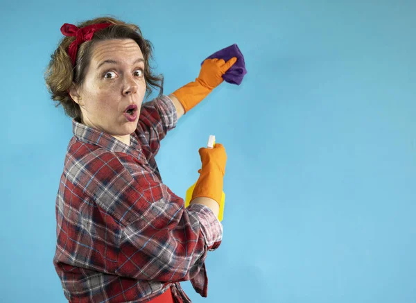 Cheerful adult woman in a plaid shirt wipes with a purple rag on a blue isolated background. Woman pin up cleaning holding a spray bottle in her hand