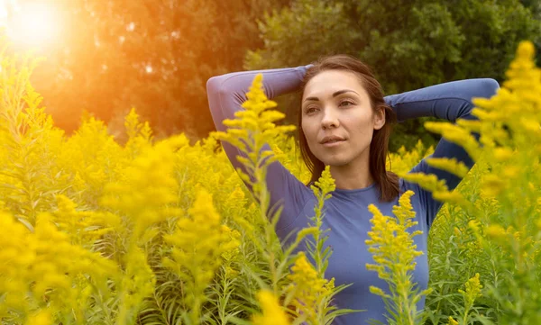 A brunette Asian woman in a blue sweater stands in yellow flowers in nature. Sunset over a woman. Asian woman holding her head with her hands