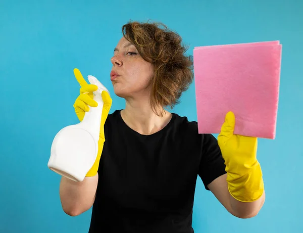 An adult cleaning woman in a black t-shirt holds a white spray bottle and a pink cleaning rag in her hands. Portrait of woman cleaning in yellow gloves