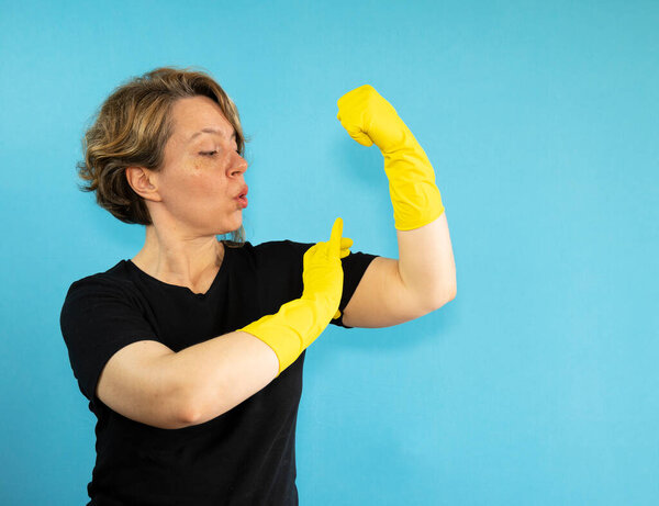 A forty-year-old cleaning woman in a black T-shirt and yellow rubber gloves shows her biceps. Portrait woman cleaning on blue isolated background