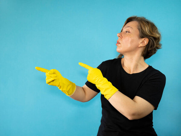 An adult woman in a black t-shirt and rubber yellow gloves shows her fingers on a blue isolated background. Woman cleaning. Portrait of a forty year old woman