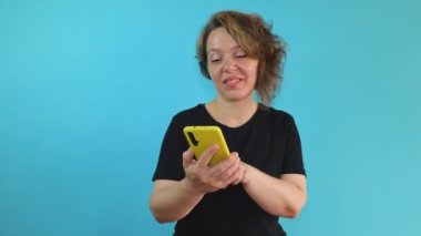 Cheerful forty-year-old woman in a black T-shirt holds a mobile phone in a yellow case in her hands on a blue isolated background. Portrait of a cheerful adult woman