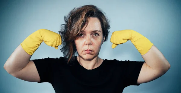 Portrait of a serious adult woman in a black t-shirt shows muscles on the background. Woman cleaning in yellow gloves. Vintage woman portrait