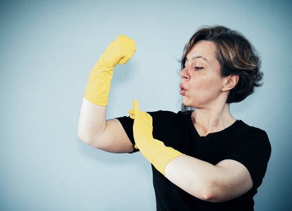 A cleaning woman in a black t-shirt looks at the biceps on the background. Vintage woman portrait. Woman in yellow rubber gloves