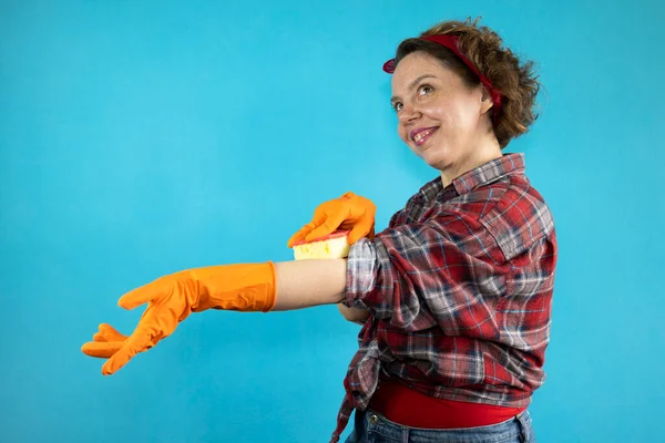 Cheerful pin-up cleaning woman in a plaid shirt wipes her body with a washing sponge on a blue isolated background. Portrait of cheerful woman cleaning in orange gloves