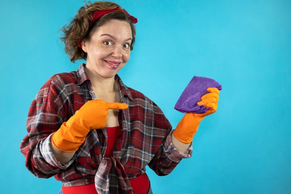 Cheerful pin-up cleaning woman in a plaid shirt holds a purple rag in her hand on a blue isolated background. Portrait of woman cleaning in orange gloves