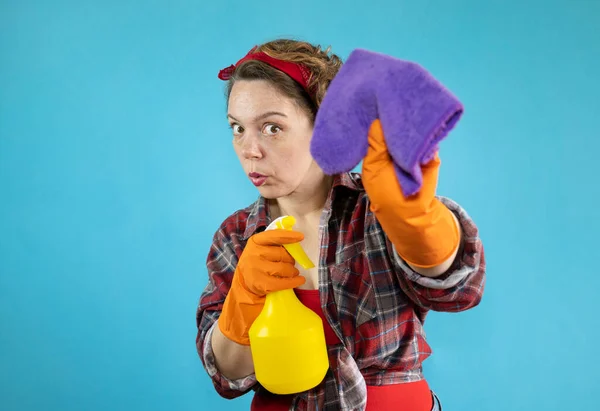 A pin-up cleaning woman with a yellow spray bottle holds a purple cleaning rag in her hand on a blue isolated background. Portrait of woman cleaning