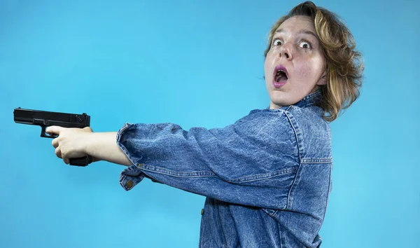 A woman with an open mouth in a denim jacket shoots a gun on an isolated blue background. American woman portrait