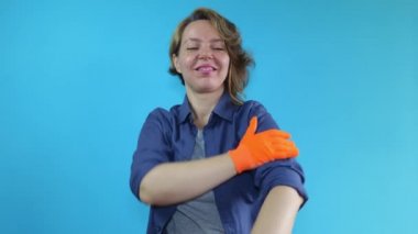 Forty year old woman in a shirt and orange gloves shows muscle strength on an isolated blue background. Portrait of a working woman. Beautiful adult woman