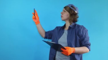 An adult woman in a shirt and a cap holds a clipboard and a pen in her hand on a blue isolated background. Woman builder shows pen towards blue background