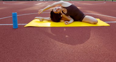 A young athletic woman in a black suit is stretching on a yellow mat in the middle of a stadium. A woman is training in the city. Portrait of a sporty woman