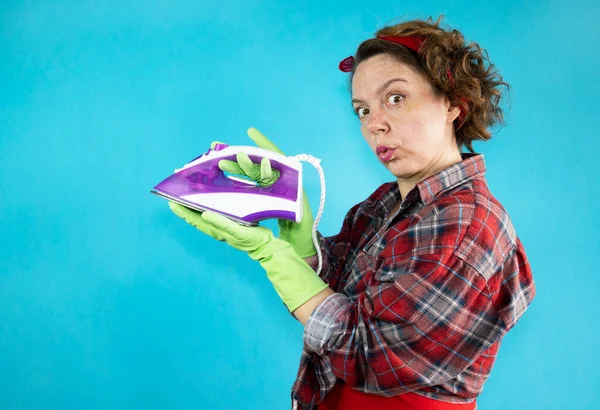 A pin-up woman in a plaid shirt and green gloves holds an iron in her hands on a blue isolated background. Woman cleaning. Portrait of a woman. Adult pin-up woman