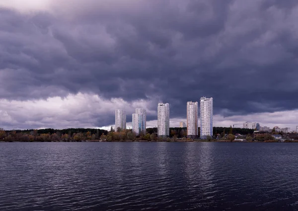 View across the dark lake to tall new houses in the city near nature. Fantasy nature. Dark cloudy landscape. Dark terrible clouds
