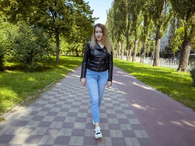 A young woman in a leather jacket and denim pants walks along the road in the city park. Woman in motion