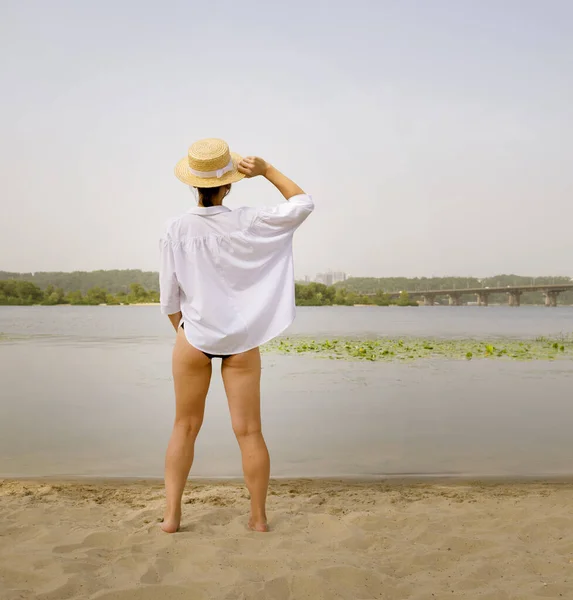 Rear view of a young woman in a hat and white shirt looks at the river and the bridge in the distance in the city. Woman on the beach near the water