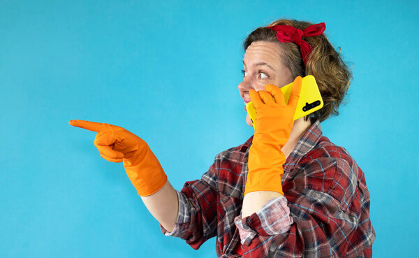 Pin-up woman in a plaid shirt talking on a mobile phone in a yellow case on a blue isolated background. Woman cleaning in orange gloves pointing finger