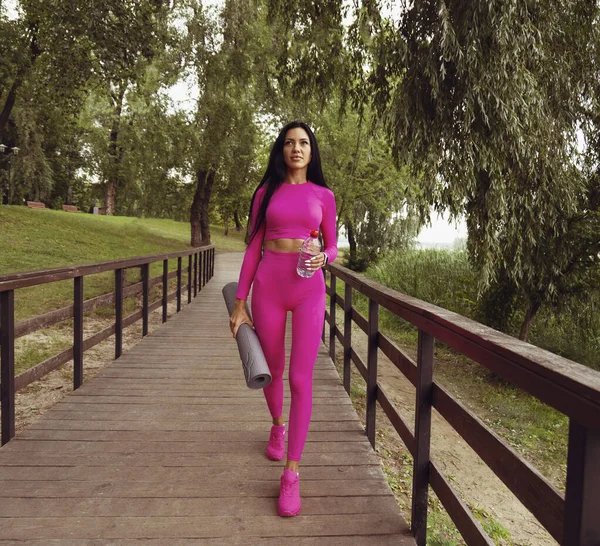 A brunette woman in a pink sports suit walks along a wooden bridge holding a sports mat and a bottle of water in her hands. brown tint