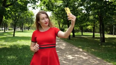 Kırmızı elbiseli genç bir kadın parkta cep telefonuyla selfie çekerek yol boyunca yürüyor. Ağır çekim portresi olan kadın.