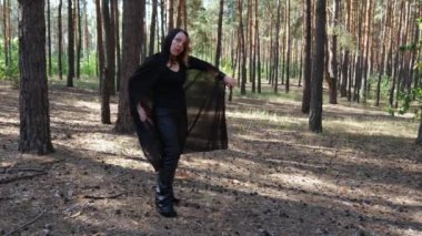 Siyah pelerinli bir kadın çam ormanında hareket ediyor. Doğadaki Cadılar Bayramı kadını. Peri ormanı. Kadın büyüsü