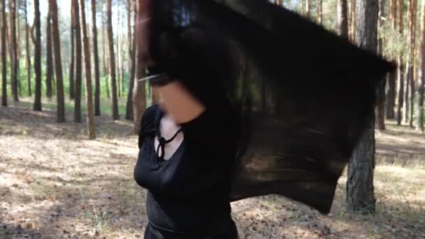 松の森の中に立っていると フード付きの黒いマントを着た大人の女性が手を振っている ハロウィーンの女性の肖像画スローモーション 女魔術師 — ストック動画