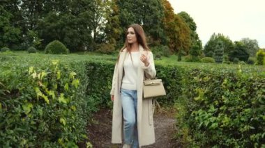 Bej ceketli genç bir kadın botanik bahçesindeki yeşil çalıların yanında yürüyor. Yavaş çekim portresi. Sonbaharda doğada yürüyen bir kadın