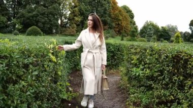 Bej ceketli esmer bir kadın botanik bahçesindeki yeşil bir çalılığın yanında yürüyor. Güzel bir kadının portresi. Sonbaharda hareket eden kadın