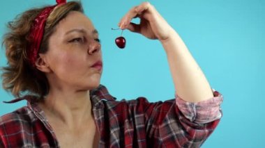Ekose gömlekli güzel, yetişkin bir kadın izole edilmiş mavi arka planda kiraz yiyor. Bir kadının yavaş hareket eden portresi. Pin-up kadın portresi