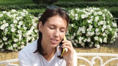 Beyaz gömlekli genç esmer bir kadın parkta bir bankta otururken cep telefonuyla iletişim kuruyor. Bir kadın portresi. Yavaş çekim portresi