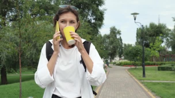 公園をバックパック付きの白いシャツを着た若いブルネットの女性が歩いている 女性が黄色いカップからコーヒーを飲んでいて 携帯電話で話している スローモーションポートレート — ストック動画