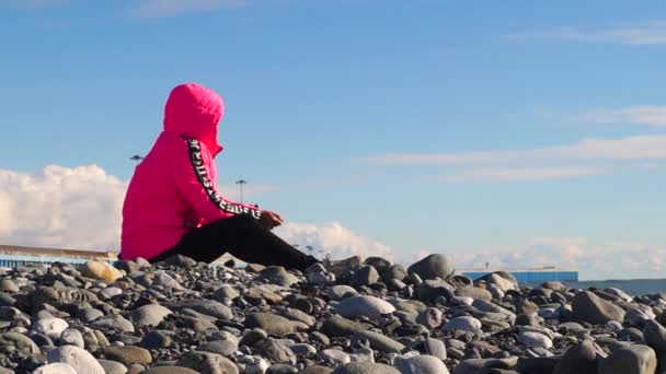 Ένα νεαρό κορίτσι με κόκκινο μπουφάν κάθεται σε μια βραχώδη παραλία και κοιτάζει μέσα στην απόσταση. — Αρχείο Βίντεο