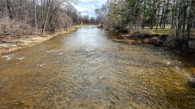 Bu nehirle birlikte kış artık donmadı. Ağaçlar hala çıplak. Bulutlu mavi gökyüzü ile aydınlık bir gün. Nehrin aşağısına uzanan bir köprü var..