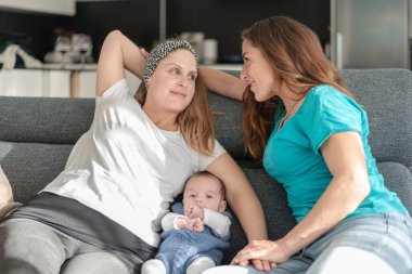 Yavru eşcinsel anne babalarıyla koltukta oturan genç bir lezbiyen çift, eşcinsel evlilik ve evlat edinme kavramı