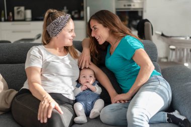 Yavru eşcinsel anne babalarıyla koltukta oturan genç bir lezbiyen çift, eşcinsel evlilik ve evlat edinme kavramı