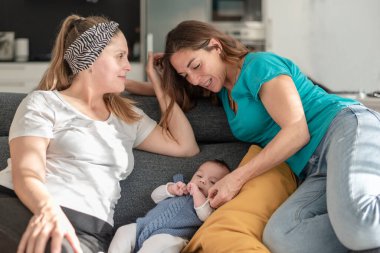 Yavru eşcinsel anne babalarıyla koltukta oturan genç bir lezbiyen çift, eşcinsel evlilik ve evlat edinme kavramı
