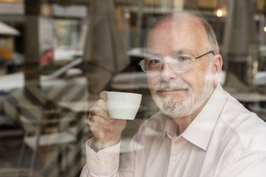 Gözlüklü, gülümseyen yaşlı bir adamın portresi. Kahve barının içinde oturmuş, elinde kahve bardağıyla pencereden dışarı bakıyor.